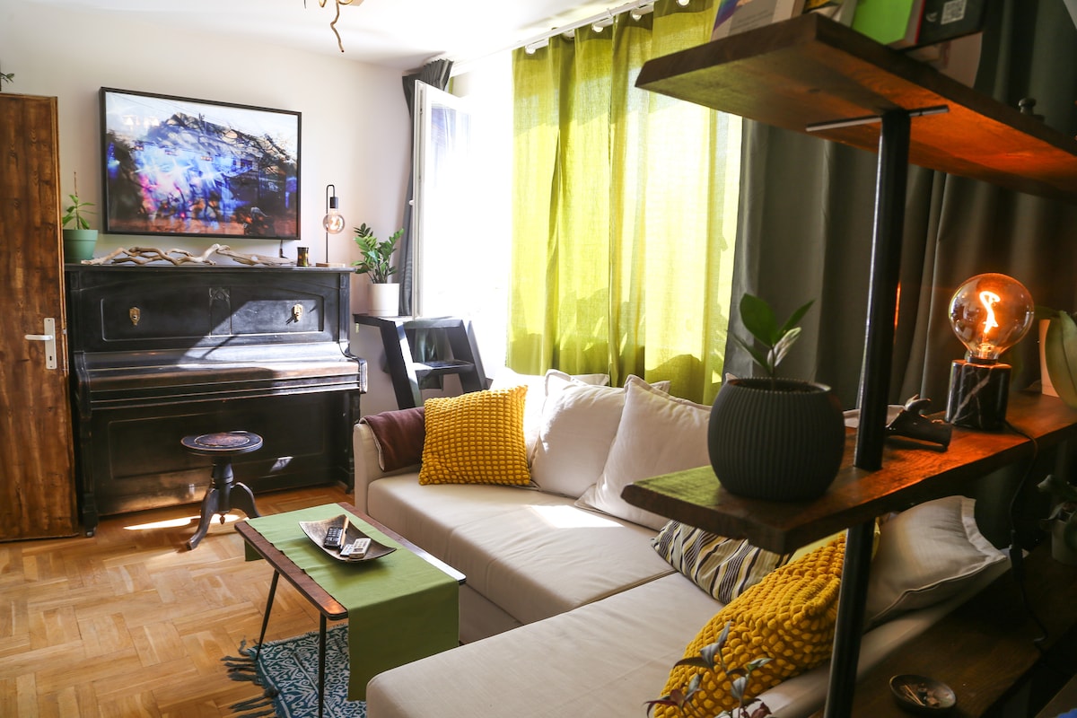 A cozy living area is highlighted by a comfortable sofa adorned with decorative cushions. An antique piano sits in the corner, complemented by natural light filtering through green curtains. A modern bookshelf features small plants and decorative items, creating an inviting atmosphere.