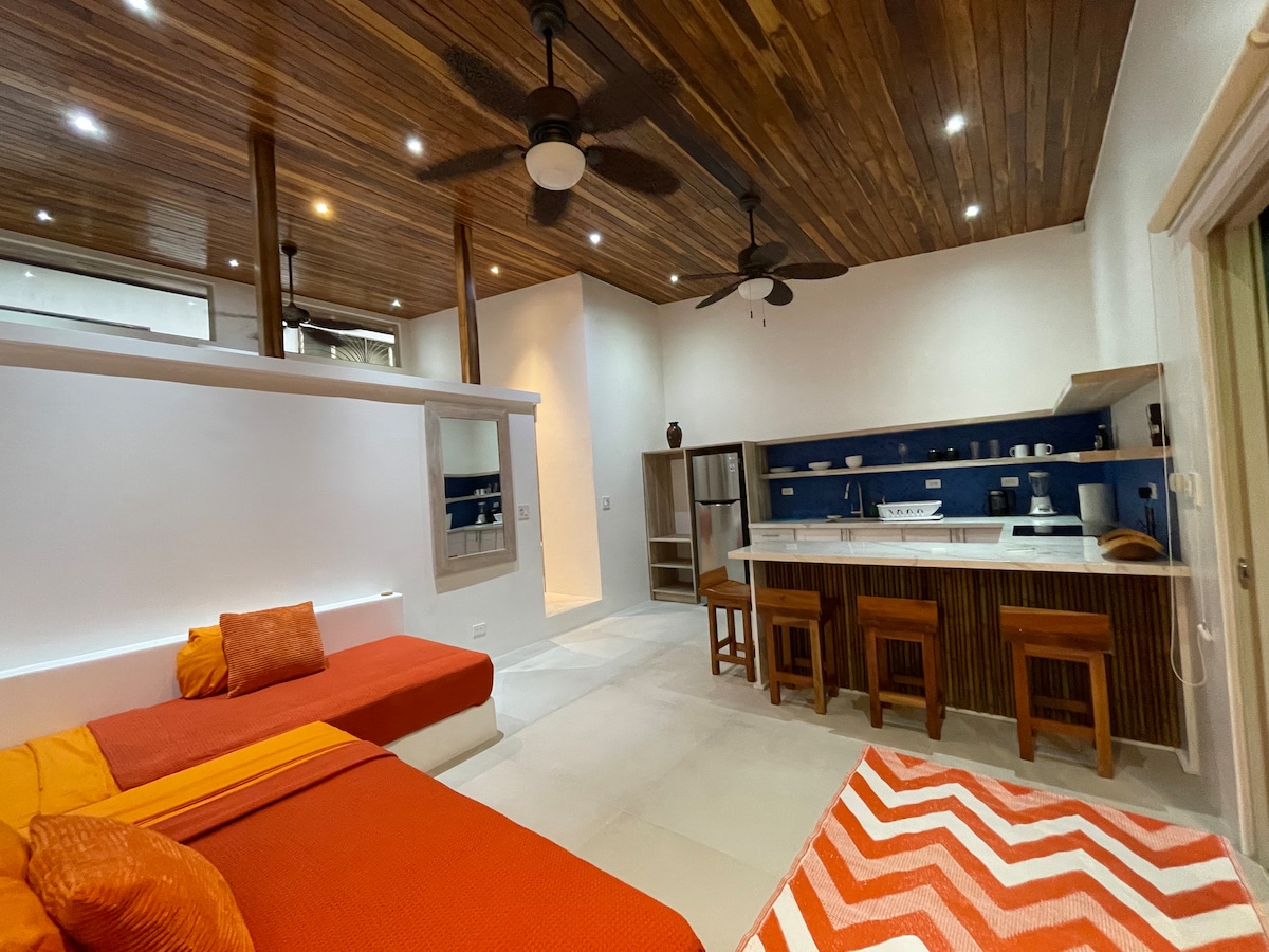 The living area features twin day beds with vibrant orange bedding, complemented by decorative pillows. A spacious kitchen island is equipped with bar stools, and a blue shelf displays handware. Natural wood beams and recessed lighting accentuate the ceiling, enhancing the cozy ambiance.