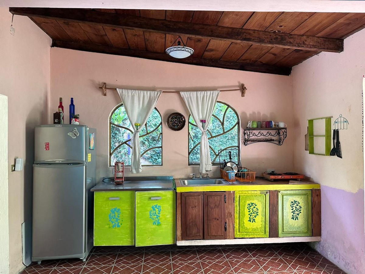 A kitchen area features a combination of bright green and wooden cabinetry. Two large windows, adorned with white curtains, allow natural light to fill the space. The refrigerator stands alongside the sink and stove, while a small shelf displays kitchen items.