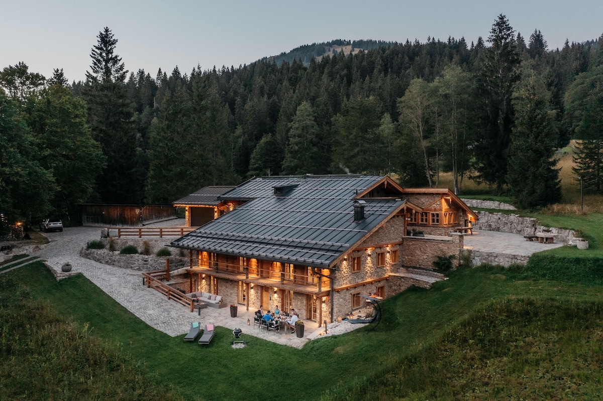 A spacious chalet is showcased, surrounded by trees and mountains. The structure features numerous large windows and a stone facade, which are illuminated warmly at dusk. An outdoor area includes seating and pathways, blending harmoniously with the natural landscape.