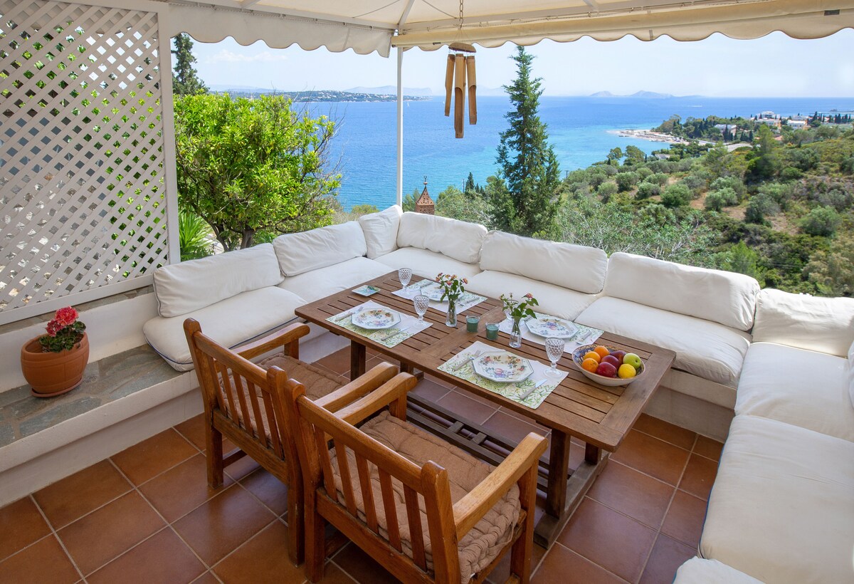 An outdoor seating area is arranged with a large sectional couch adorned with white cushions and a wooden table at the center. A variety of fruits and floral arrangements are placed on the table, with scenic views of the sea and greenery visible in the background.