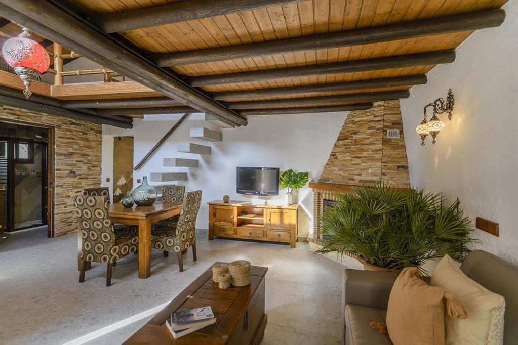 An inviting living area is highlighted by wooden beams above and a stone accent wall. A round dining table with patterned chairs sits beside a wooden cabinet. A television is mounted on the wall, and a green plant brings a touch of nature to the space.