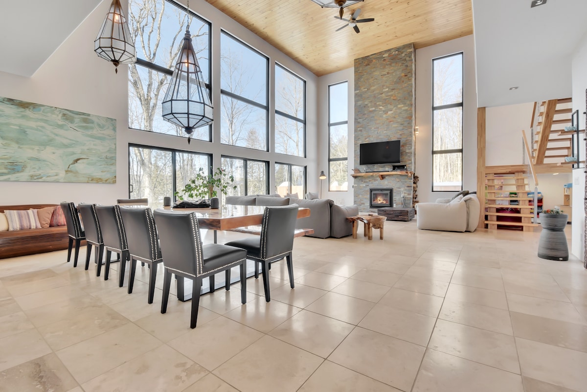 A spacious living area features large windows that allow natural light to fill the room. A modern stone fireplace is centered on a feature wall. A long dining table with black chairs is positioned in the foreground, while cozy seating areas are arranged nearby.
