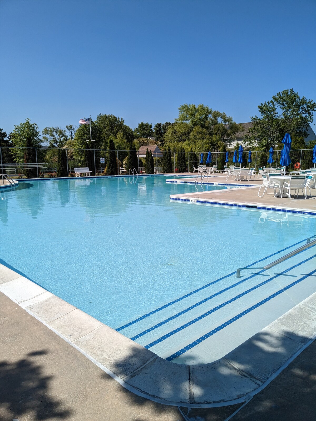 A spacious community pool is shown, featuring clear blue water reflecting the sky. Poolside lounge chairs with blue umbrellas surround the area, while a few trees provide a green backdrop. Steps lead into the inviting water, enhancing the relaxing atmosphere.