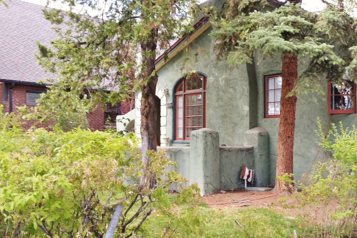 A charming exterior of a house is surrounded by lush greenery, featuring a textured green facade and arched windows. The unique design is complemented by tall trees, creating a serene and inviting atmosphere. The carefully maintained landscape adds to the character of the home.