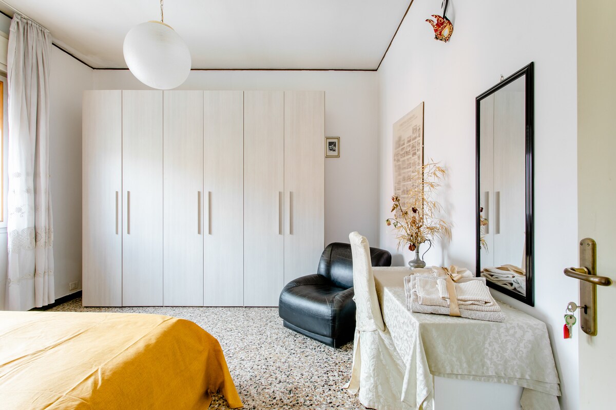 A spacious bedroom is presented with a large wardrobe featuring minimalist handles. A comfortable black chair is situated next to a desk adorned with a decorative tablecloth. Natural light filters through sheer curtains, illuminating the soft yellow bedding on the bed.