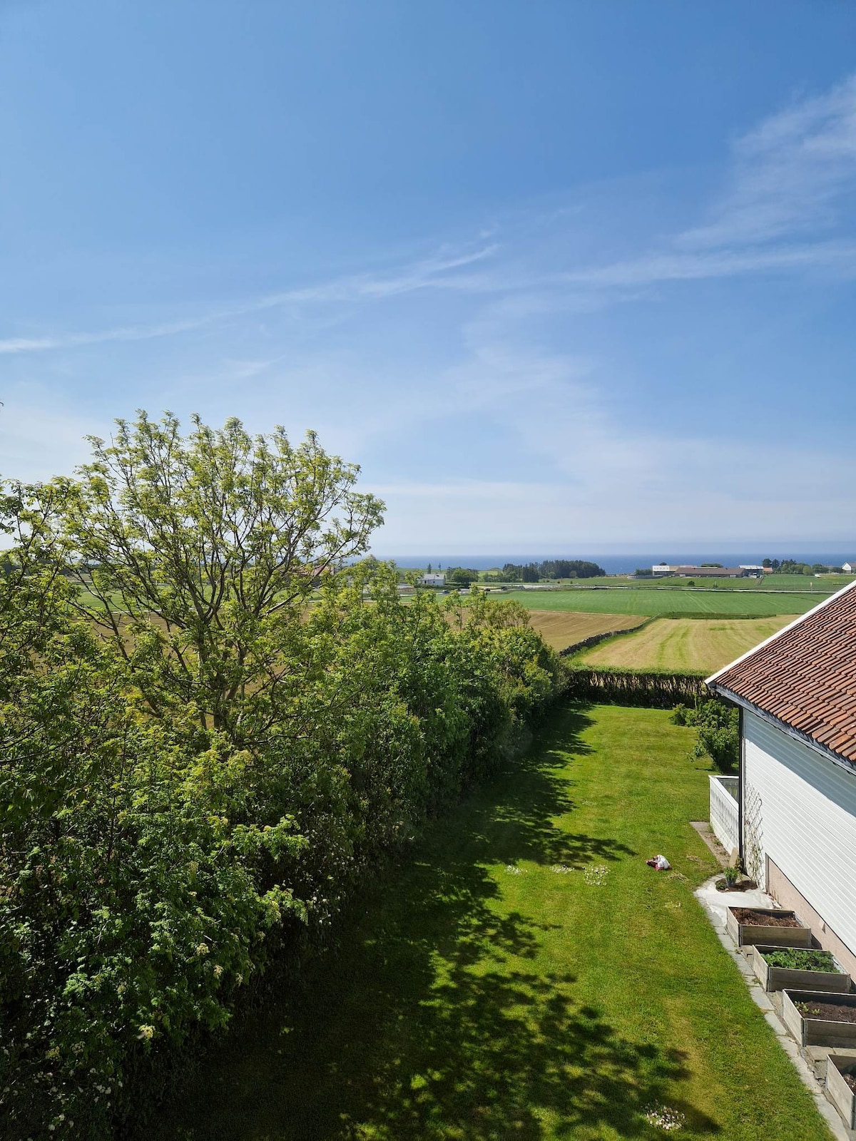 A view from the property showcases a lush green lawn bordered by trees, with a distant glimpse of the sea. The expansive sky highlights a serene landscape, while neatly kept fields extend into the horizon, providing a tranquil backdrop.