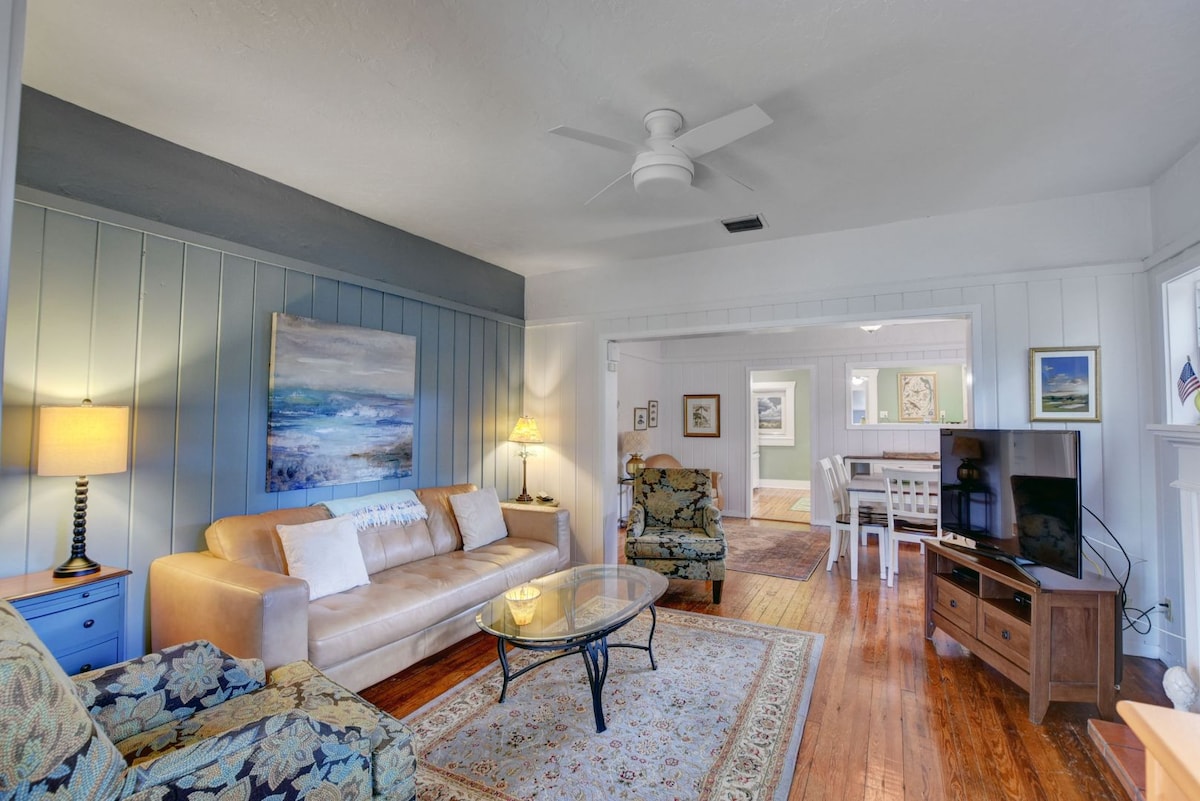 A comfortable living room features wood flooring and light-colored walls. A large painting of a serene landscape adorns one wall. A brown sofa, patterned armchairs, and a glass coffee table create a welcoming seating arrangement. Soft lighting adds a cozy feel to the space.