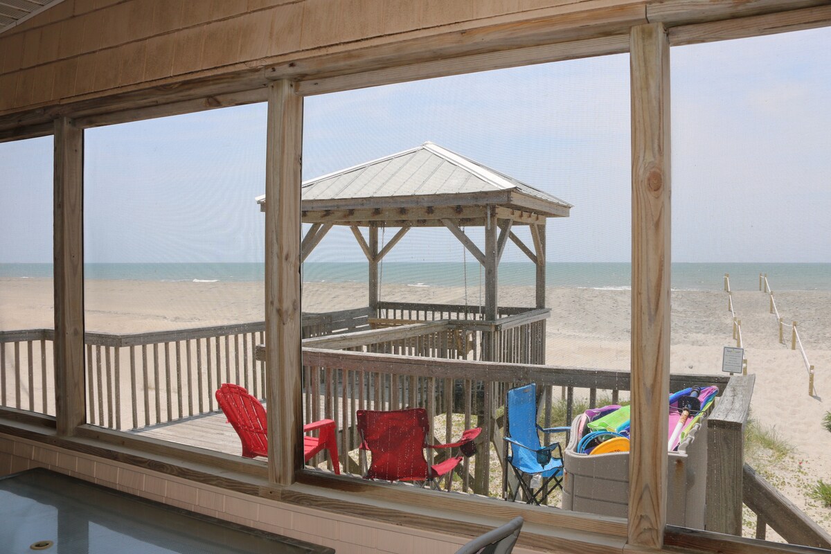 The image captures a view from the oceanfront screened-in porch showcasing a covered gazebo with a porch swing. Beach chairs in various colors are arranged nearby, with the sandy beach and water visible in the background through the screened windows.