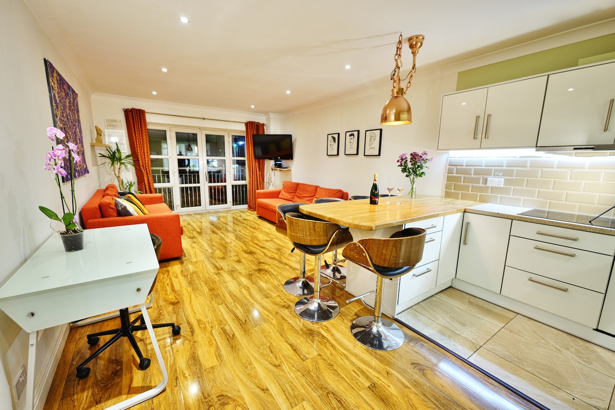 A spacious living area features a vibrant orange sofa set and a sleek kitchen with stylish fixtures. A wooden dining table with modern seating is complemented by a television mounted on the wall. Natural light fills the room through large sliding doors, enhancing the contemporary design.