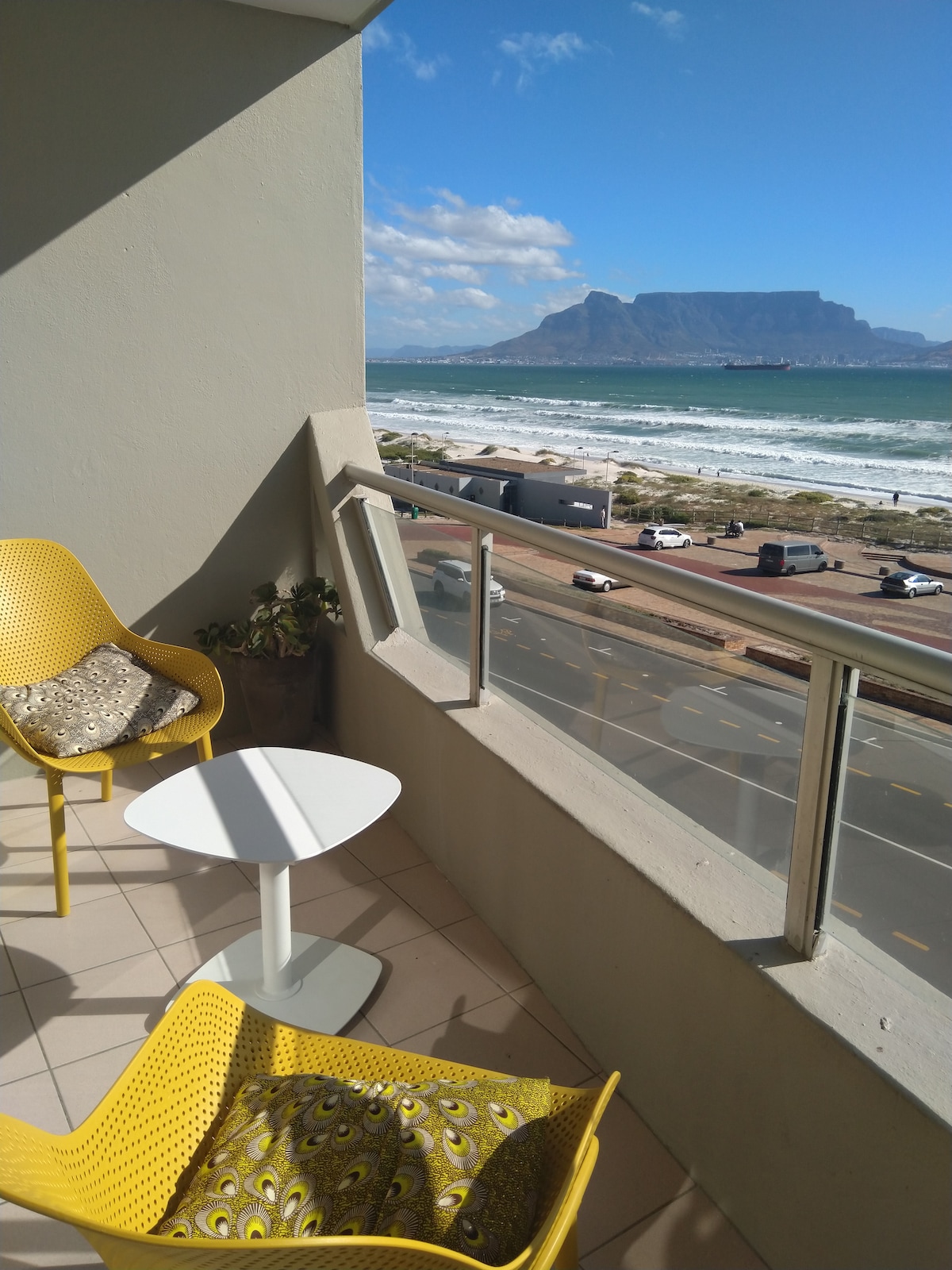 A private patio offers stunning views of the ocean and Table Mountain. Two yellow chairs with patterned cushions surround a small white table, creating a space for relaxation. The glass railing provides unobstructed sightlines to the beach and the vibrant sky.