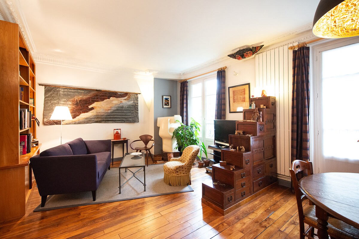 A spacious living area features a mix of vintage and modern furnishings, including a dark purple sofa, a wooden bookcase, and a decorative armchair. Large windows allow natural light to fill the room, highlighting a green indoor plant and a light-colored area rug that complements the wooden flooring.