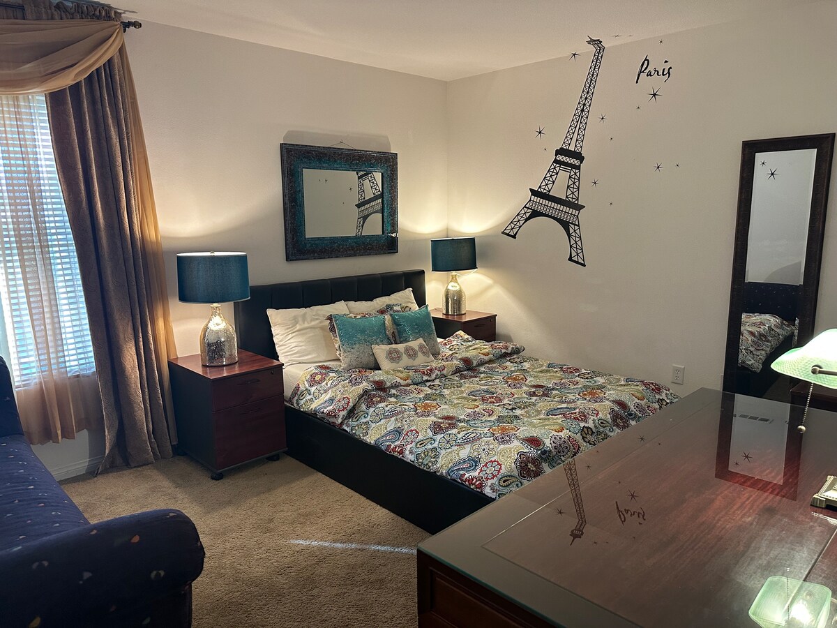 A cozy bedroom features a spacious bed adorned with a patterned quilt and decorative pillows. Two bedside tables with lamps provide soft lighting. A decorative mirror and a wall graphic of the Eiffel Tower add character to the room, while a dresser reflects the inviting ambiance.