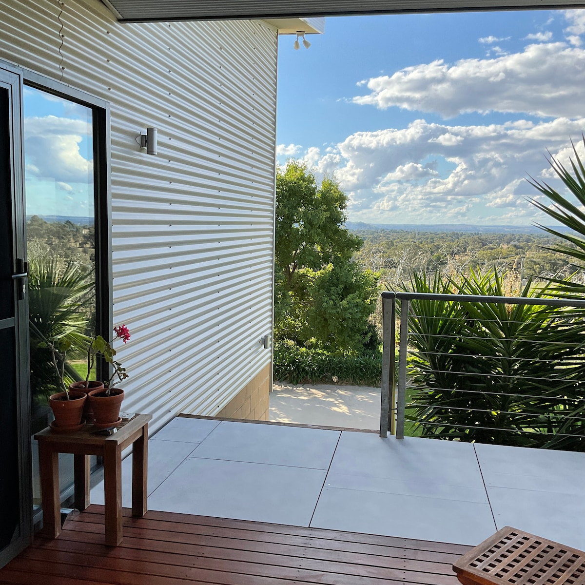 A private outdoor patio features potted plants, tile flooring, and wooden decking. Expansive views of the countryside are visible, showcasing distant trees and rolling landscapes. Natural light brightens the space, creating an inviting area for relaxation or outdoor dining.