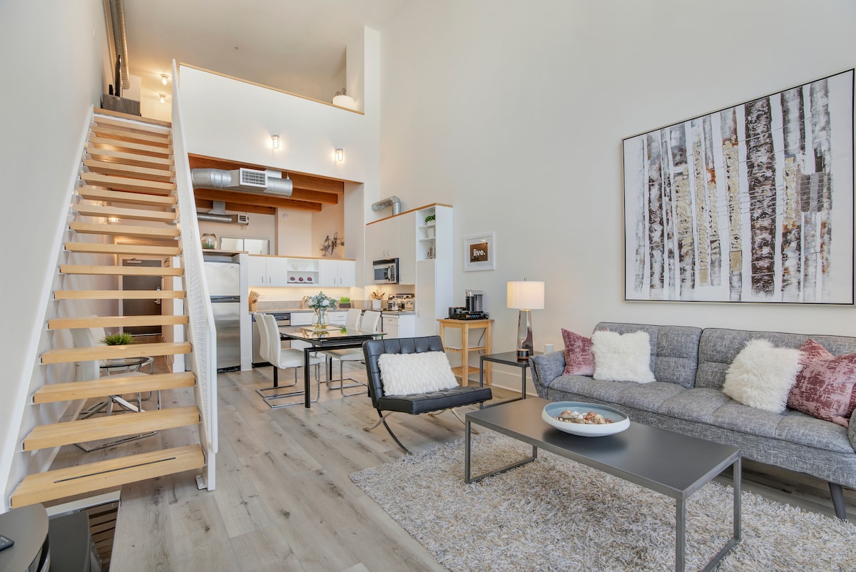 A stylish loft space features a light-filled living area with a plush gray couch and a modern coffee table. A wooden staircase leads to the upper level, and a dining area is visible in the background, complemented by an open kitchen with sleek appliances.