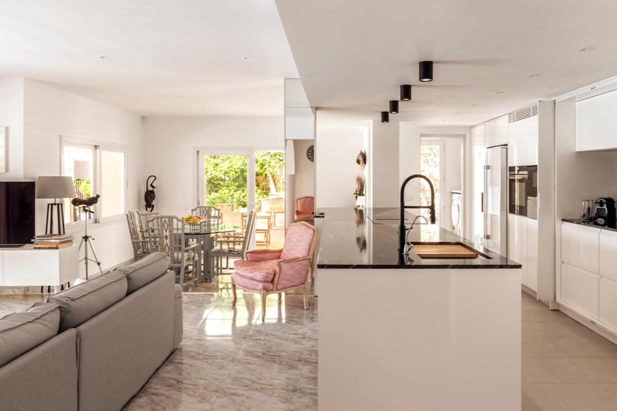An open-plan kitchen and living area features a modern design with sleek white cabinetry and a dark countertop. Natural light floods the space through large windows. A dining table surrounded by colorful chairs complements a cozy seating arrangement with a gray sofa and pink armchairs.