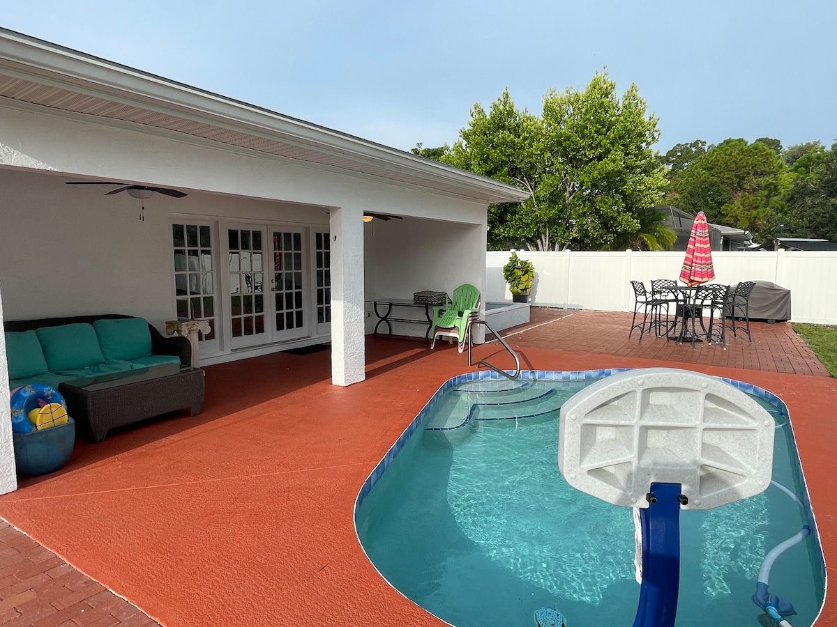 A private solar-heated pool is framed by a spacious patio, featuring a mix of lounge and dining furniture. Green chairs and a table are placed nearby, while a shaded area offers a cozy seating arrangement. A white fence provides privacy, enhancing the outdoor space.