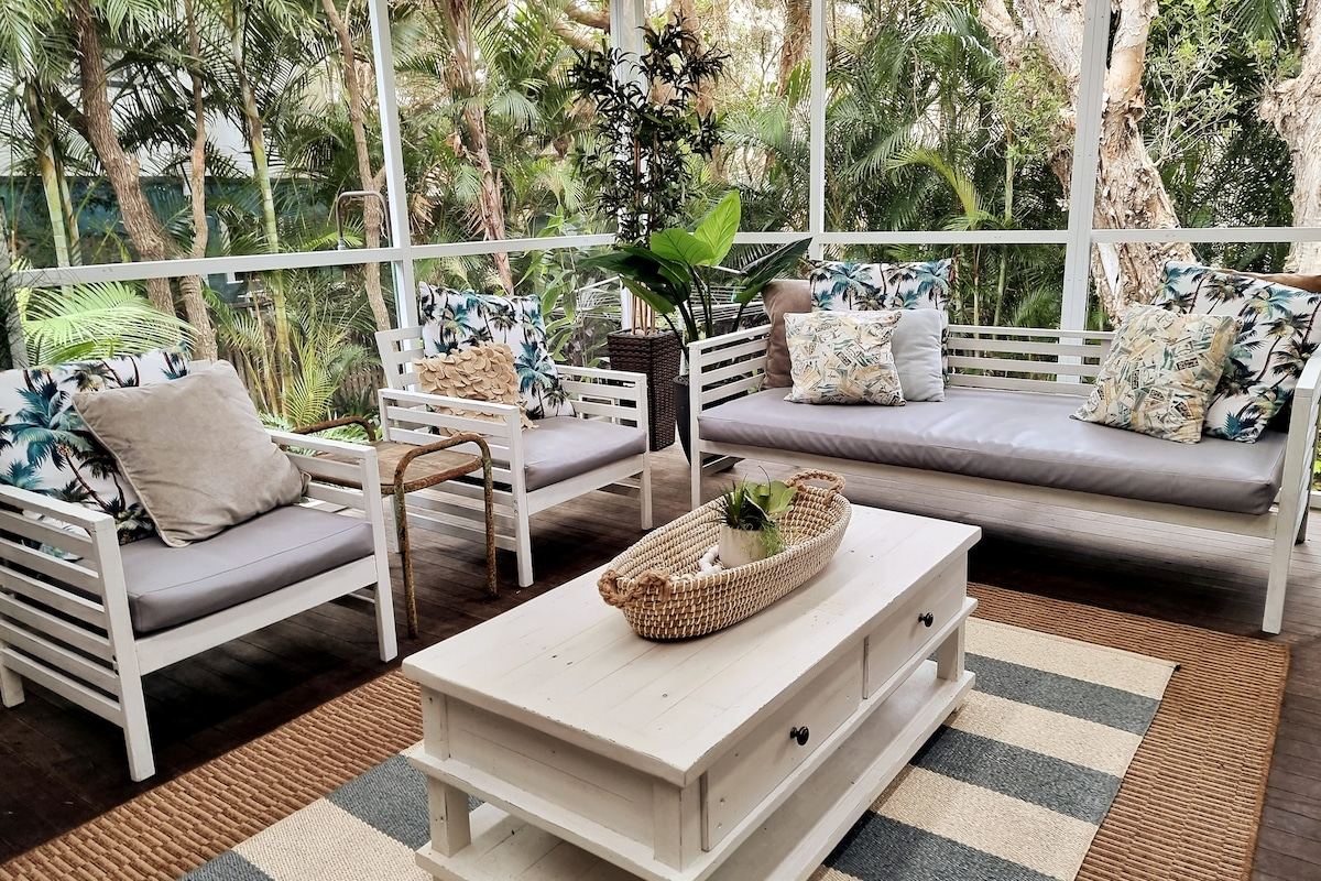 A spacious screened outdoor room features a white wooden seating arrangement with cushions in tropical patterns. A neutral coffee table is centered on a striped rug, surrounded by potted plants and foliage, creating a serene and relaxing environment.