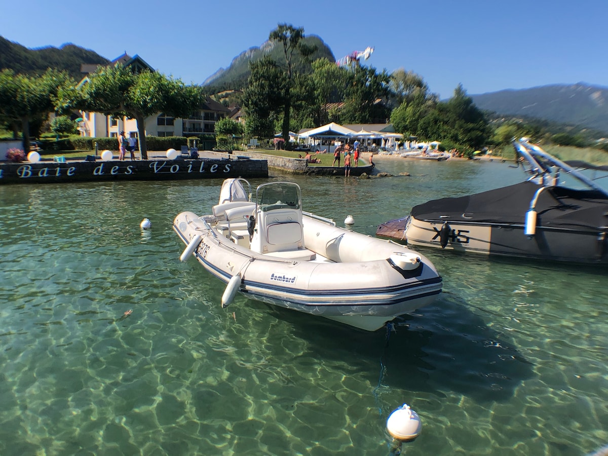 A small boat floats on clear waters of the lake, with the shoreline visible in the background. Lush greenery and a few structures line the water's edge, creating a serene lakeside scene. Mountains can be seen in the distance, under a bright blue sky.
