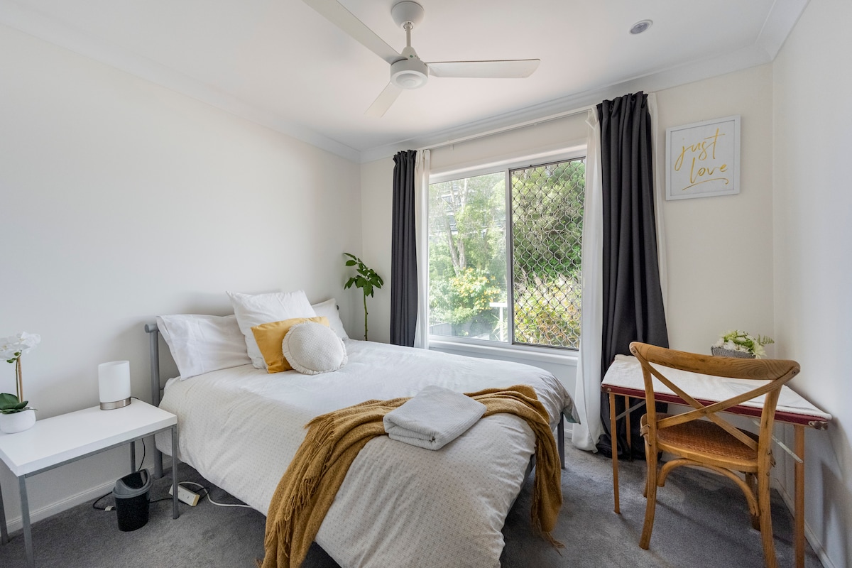 A bright bedroom features a spacious bed adorned with neutral linens and a soft throw. A desk with a chair is situated near a window framed by dark curtains, allowing natural light to filter through. A potted plant adds a touch of greenery to the space.