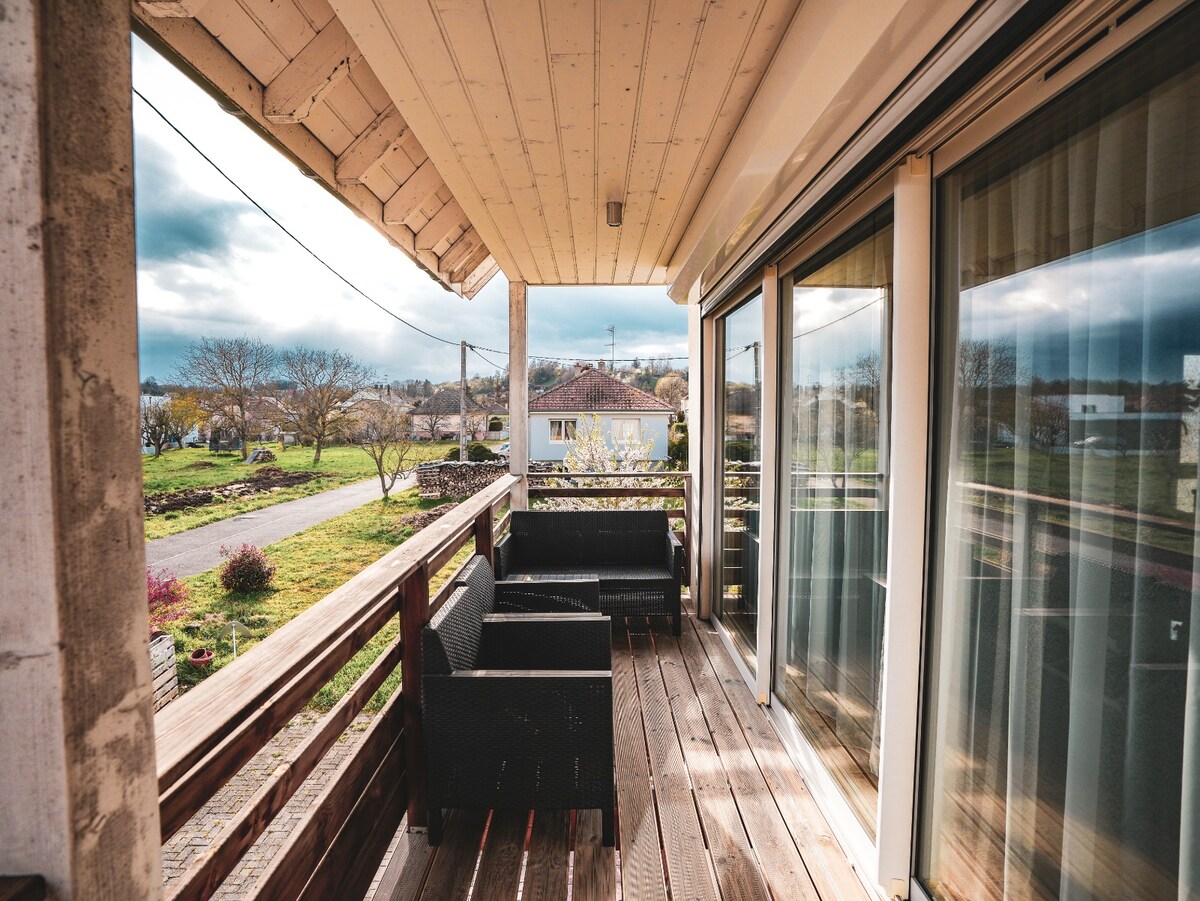 An outdoor balcony area features a wooden deck complemented by comfortable seating arrangements. Large glass doors provide natural light and a view of the surrounding greenery, enhancing the welcoming space. The scenery includes open fields and nearby residences in the distance.