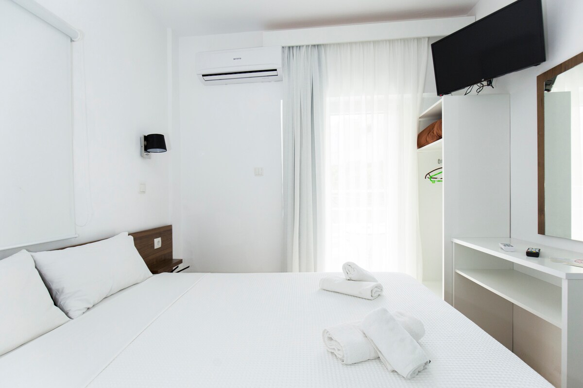 A bright room is presented, featuring a spacious bed with white linens and folded towels. A flat-screen TV is mounted on the wall, alongside a mirror and a wardrobe. Soft natural light filters through sheer curtains, enhancing the overall tranquility of the space.