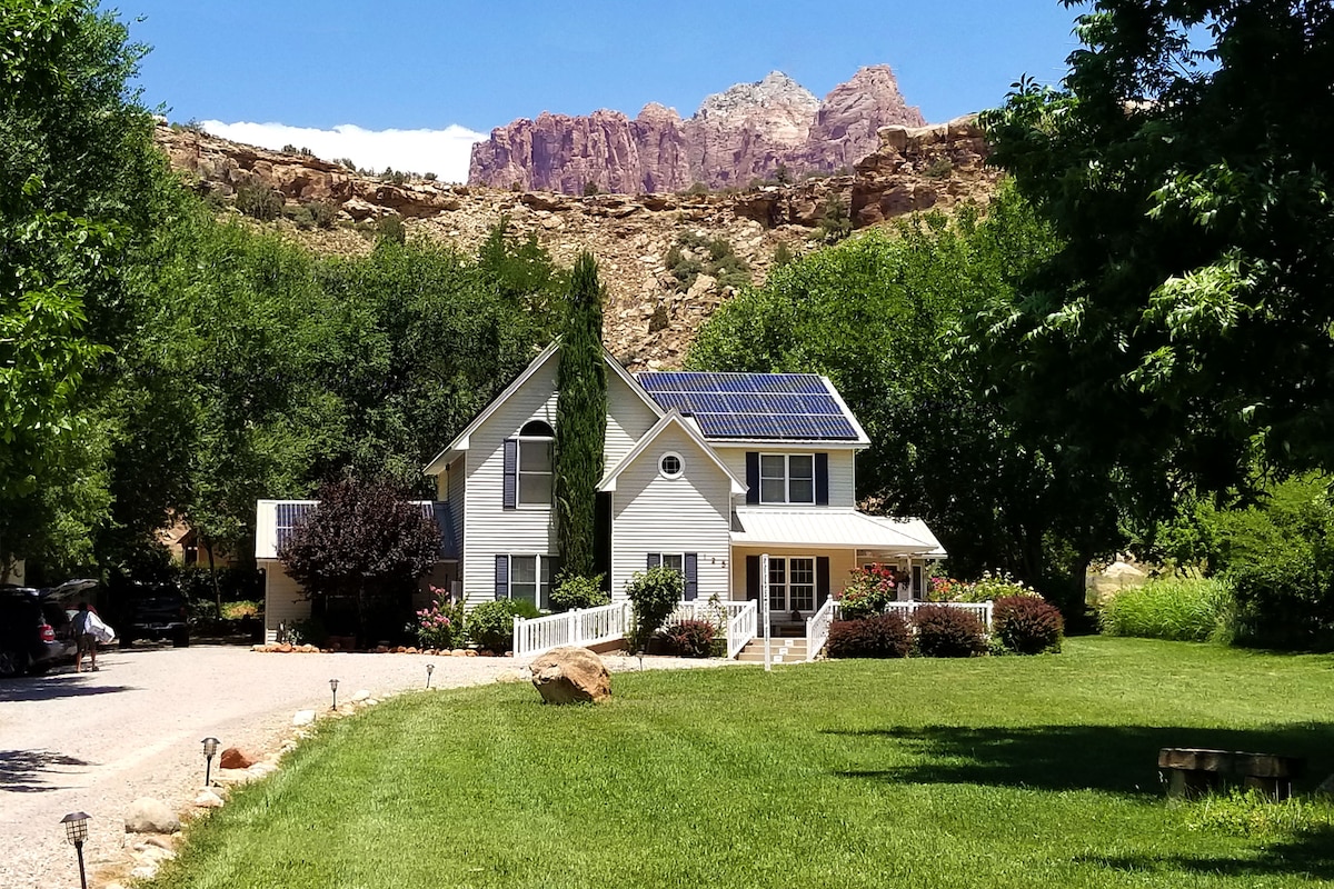 The two-story inn is positioned amidst lush greenery and features a welcoming front porch. Solar panels are visible on the roof, with picturesque canyon cliffs in the background, completing a serene outdoor setting.