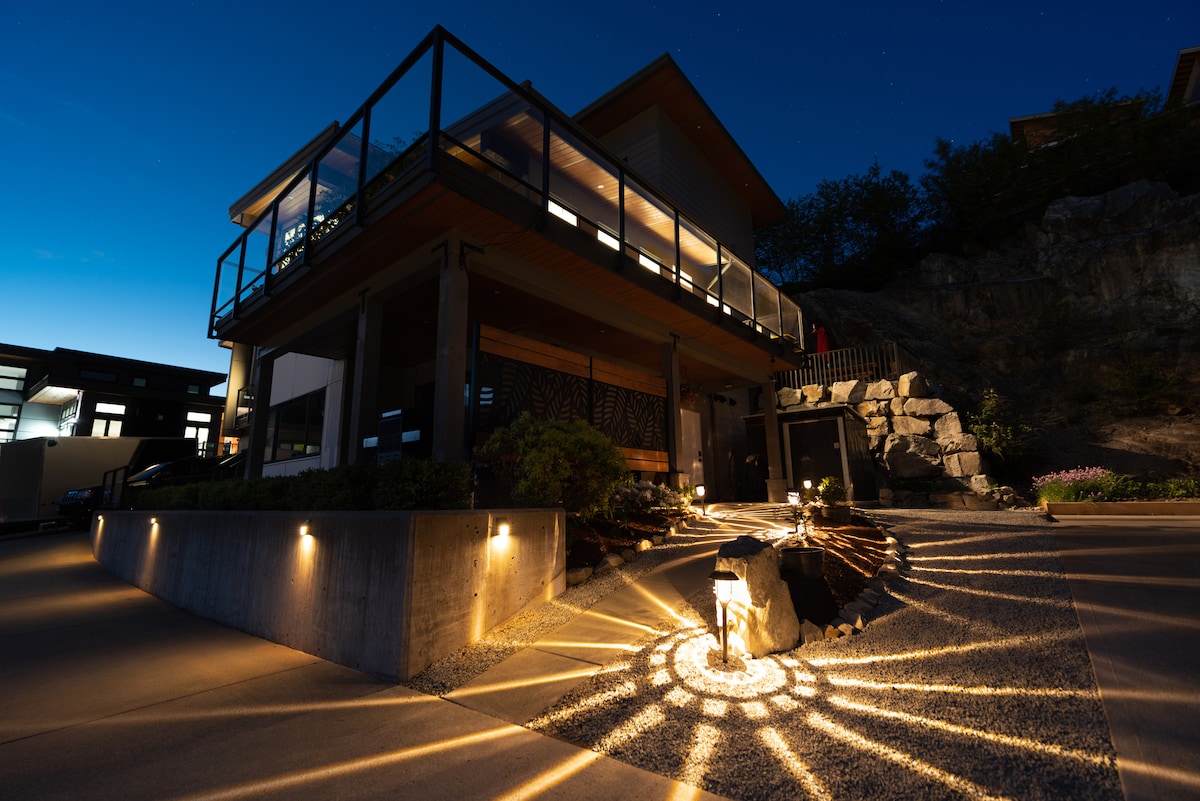 The exterior of a modern home is illuminated at night, showcasing carefully designed lighting that casts decorative shadows on the ground. A mix of natural stone and landscaping elements surrounds the entrance, enhancing the inviting presence of the residence.