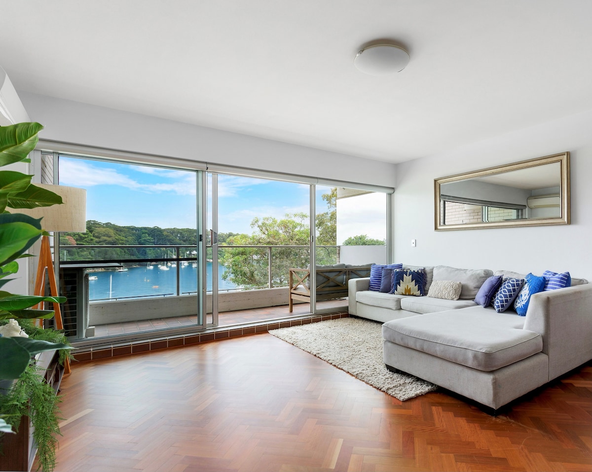 A bright living area features a large, comfortable sectional sofa enriched with decorative cushions. Sliding glass doors open to a balcony overlooking Berry's Bay, while a sunlit rug complements the warm wooden flooring. A floor lamp and mirror add functional elegance to the space.
