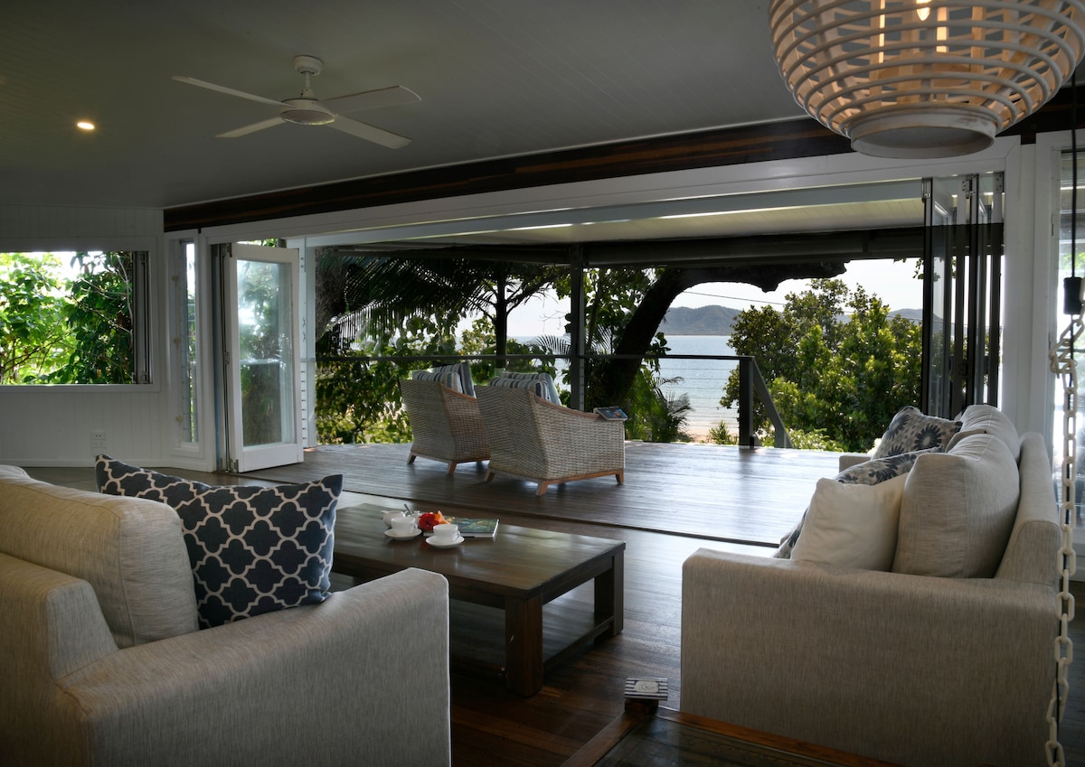 A spacious living area is highlighted by comfortable seating arrangements, facing large glass doors that open to a scenic outdoor space. The calming view of palm trees and the ocean can be seen in the background, enhancing the sense of relaxation.