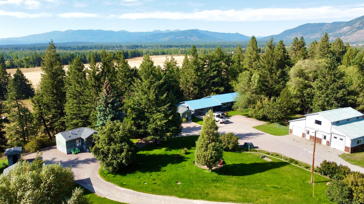 An aerial view showcases the cabin and surrounding greenery, with lush trees and open fields extending to the mountains in the distance. The property features well-maintained landscaping, a driveway, and outdoor spaces, inviting exploration and relaxation in a peaceful natural setting.