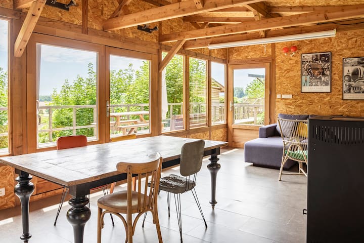 A spacious common area is illuminated by natural light through large windows, showcasing views of greenery. A dark wooden dining table is accompanied by a mix of chairs, while a cozy seating area includes a sofa and stylish accent chairs, creating a welcoming space.