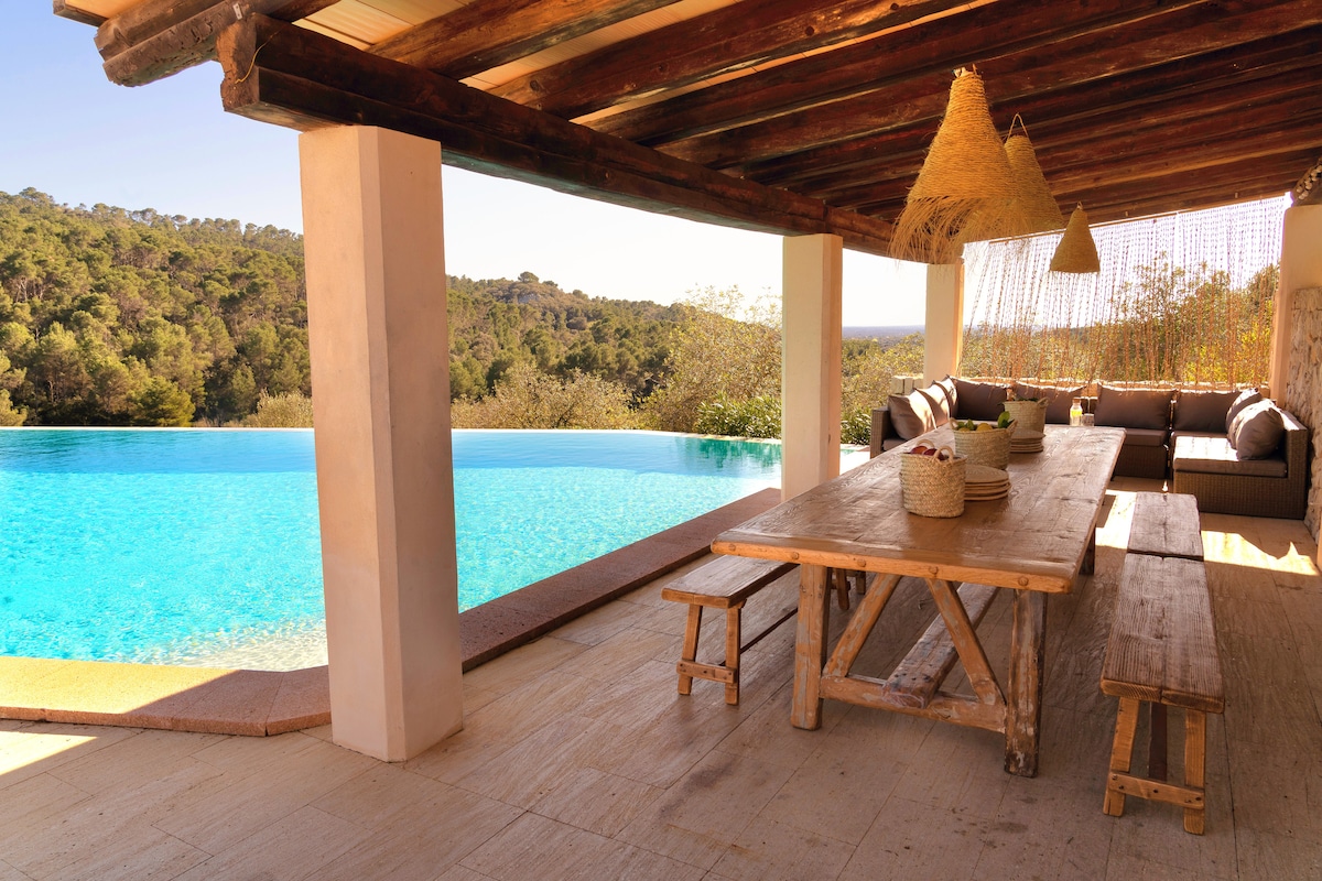 A spacious outdoor dining area features a rustic wooden table with seating alongside a comfortable lounge space. Natural fibers adorn the ceiling, and views of a large infinity pool blend with trees in the background, creating a peaceful setting for relaxation.