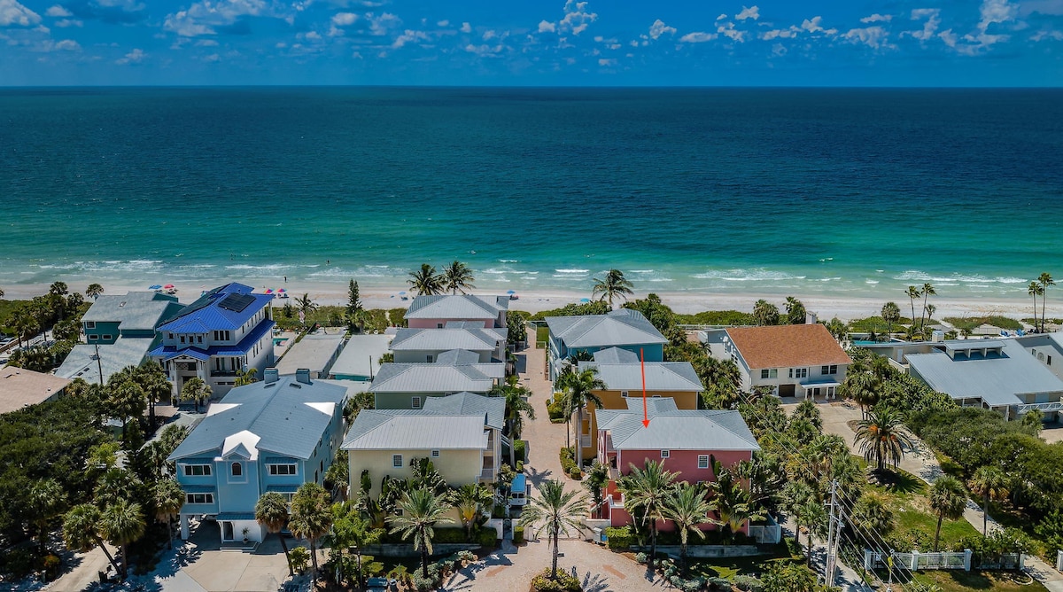 An aerial view showcases the Westwinds Paradise Beach House amidst a serene gated community. The vibrant colors of the homes contrast with the turquoise waters of the Gulf of Mexico, with sandy beaches visible in the distance under a bright blue sky.