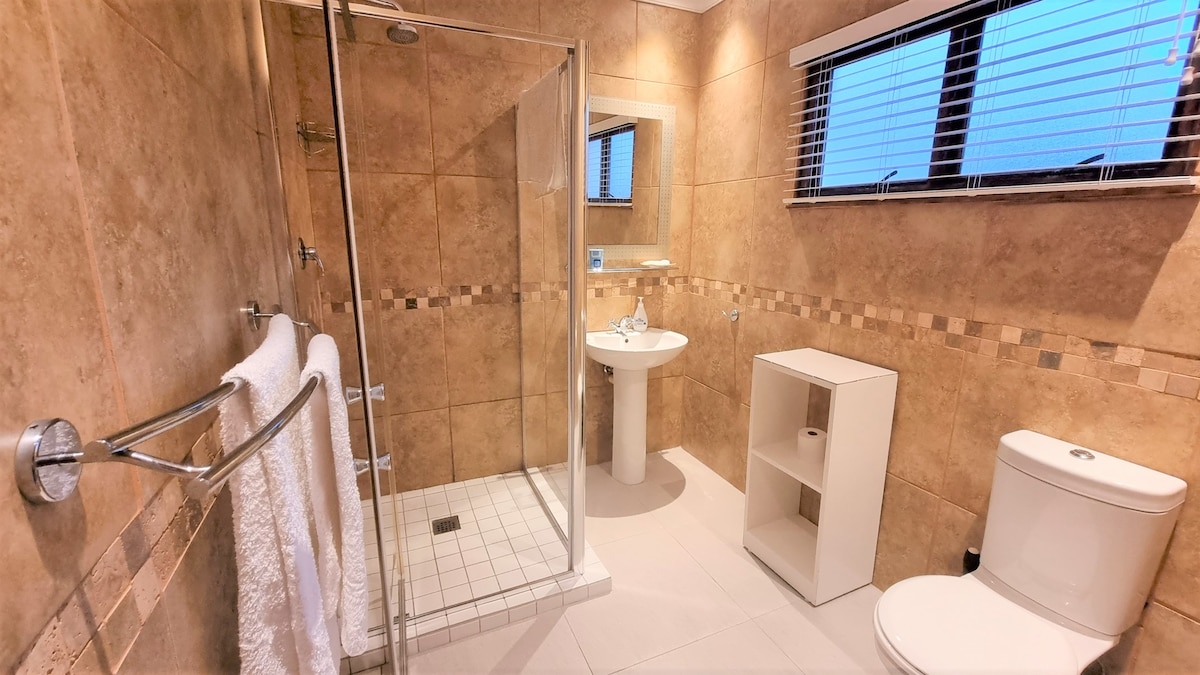 A modern bathroom features a glass shower enclosure, a white toilet, and a pedestal sink. The walls are adorned with neutral tiles, complemented by a light-colored tile floor. Natural light enters through window blinds, enhancing the spacious feel of the room.