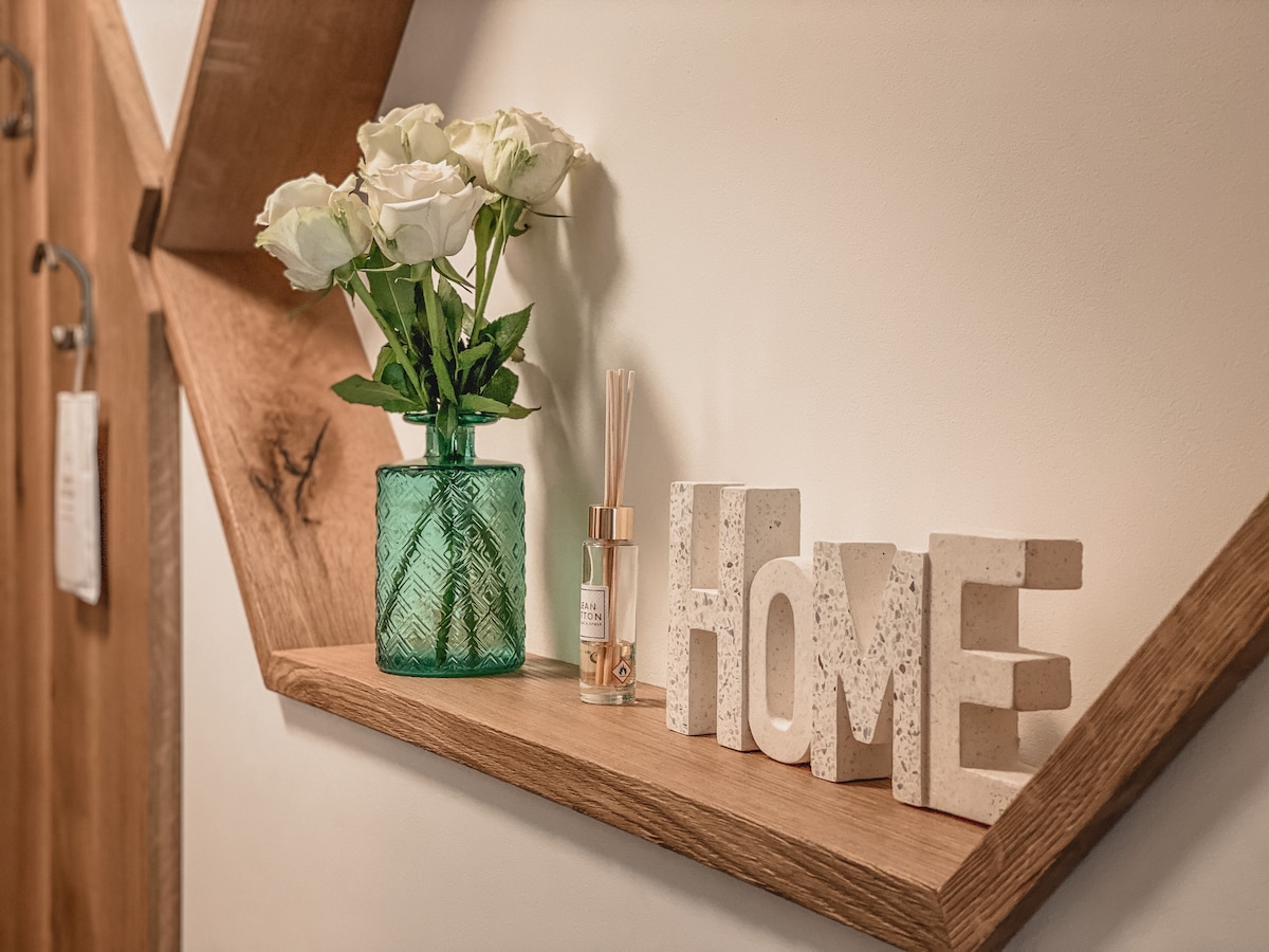 An arrangement of fresh white roses in a green glass vase sits on a wooden shelf. Decorative letters spelling 'HOME' are displayed nearby, alongside a diffuser for fragrance. The warm wooden tones of the shelf add a cozy touch to the space.