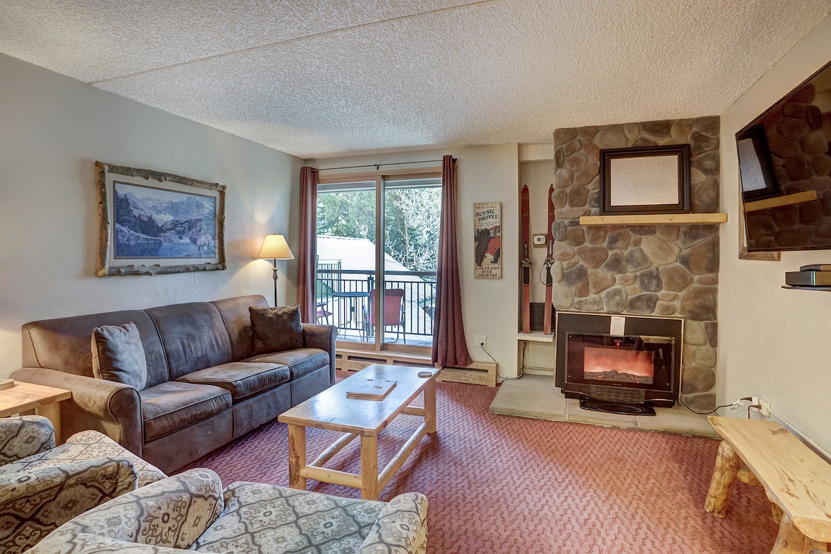 The living room features a comfortable couch and two patterned armchairs arranged around a wooden coffee table. A stone fireplace is positioned next to a mounted television. Natural light enters through sliding glass doors that lead to a small outdoor area.