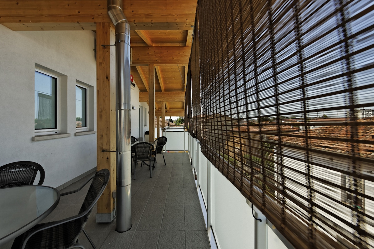 A covered terrace area features a mix of natural wood and modern materials. Black wicker chairs surround a glass table, while a woven bamboo screen provides shade. The space exudes a relaxed atmosphere with views of the surroundings visible beyond the railing.