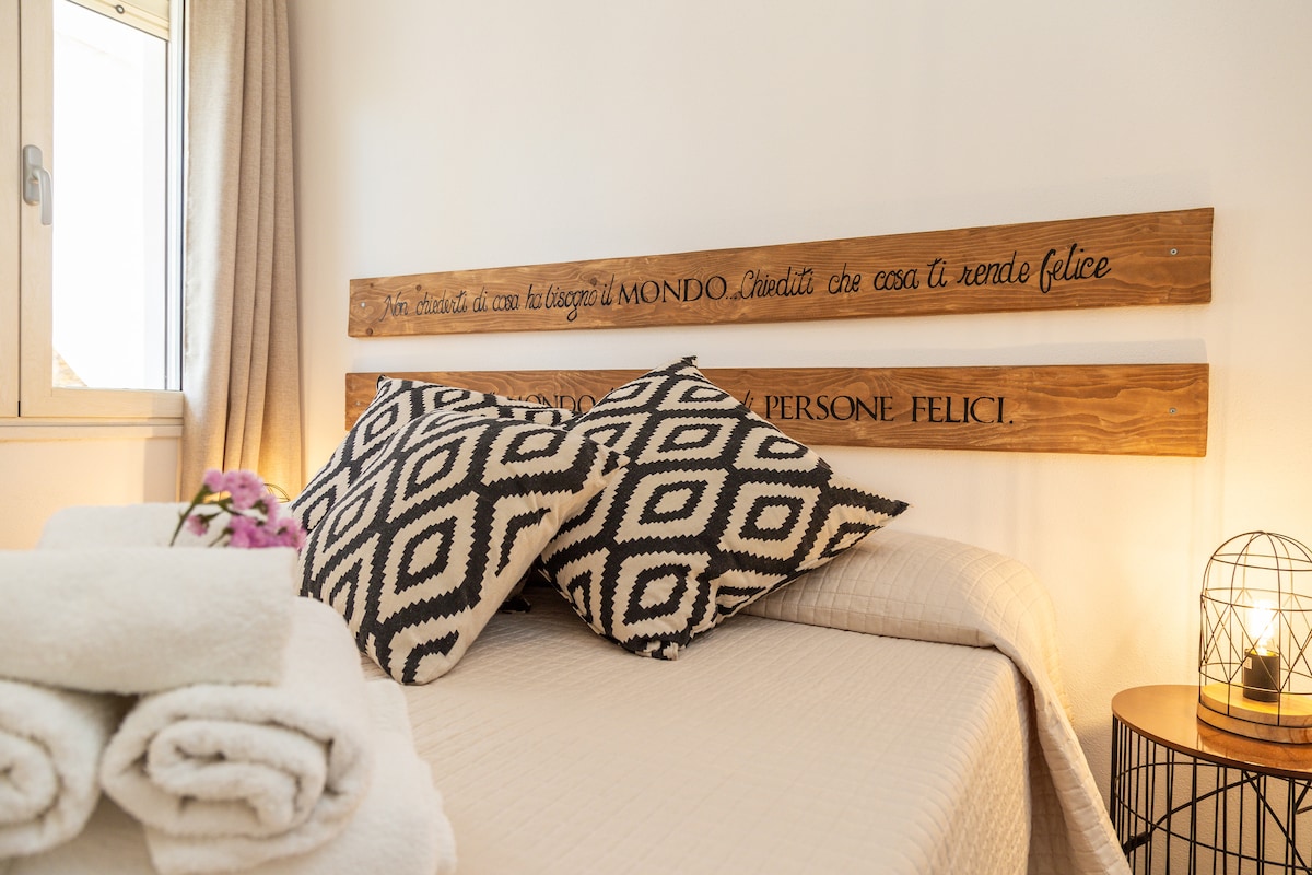 A well-appointed sleeping area features a wooden headboard with an inscription. The bed is adorned with patterned cushions and a light quilt. Nearby, stacked towels provide convenience, while a softly glowing lantern adds a touch of warmth to the space.