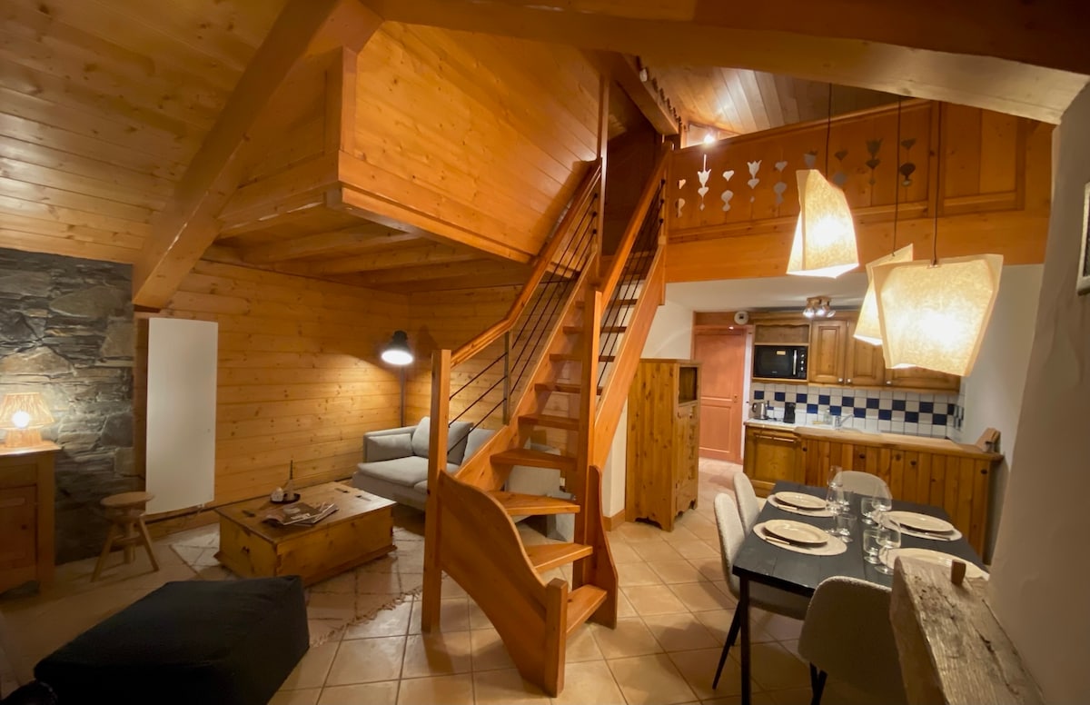 The interior features wooden walls and ceilings, creating a warm ambiance. A staircase leads to an upper level, while an inviting living area includes a soft couch and a wooden coffee table. Overhead, pendant lights illuminate the dining area with a rustic table and wooden chairs.
