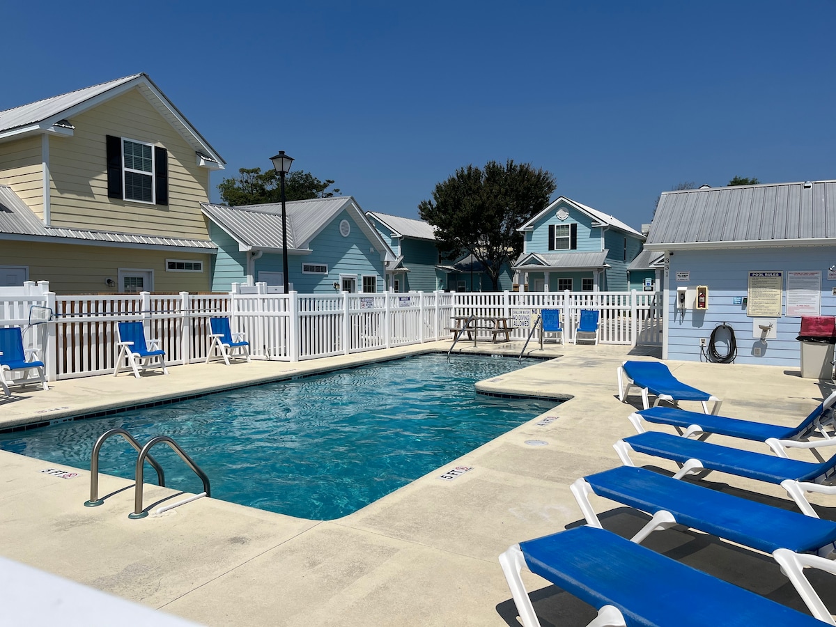 An outdoor community swimming pool is surrounded by bright lounge chairs and a privacy fence. Clear blue water reflects the sunny sky, while several cottages with pastel-colored exteriors can be seen in the background, contributing to a relaxed atmosphere.
