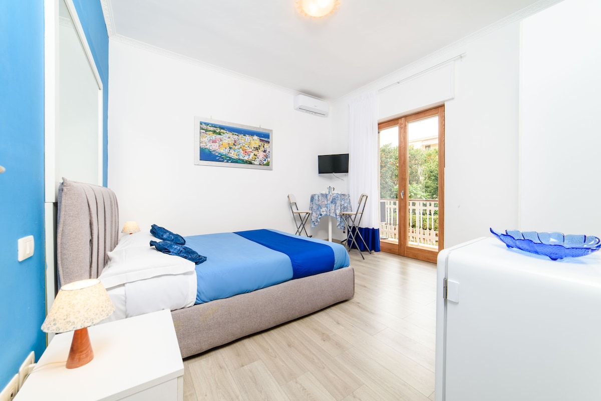 A bright studio room features a spacious bed with blue bedding against a light wall. Two windows open to a small balcony overlooking greenery. A compact kitchenette and a small dining table with two chairs are present, accompanied by a white refrigerator and flat-screen TV mounted on the wall.