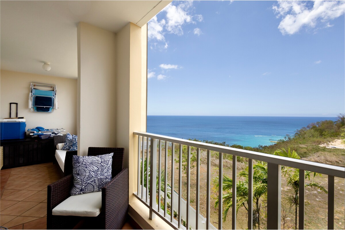 A spacious balcony offers stunning sea views, with comfortable seating arranged thoughtfully. Lush greenery is visible in the foreground, complementing the serene blue waters. The horizon is clear, enhancing the refreshing outdoor space perfect for relaxation.
