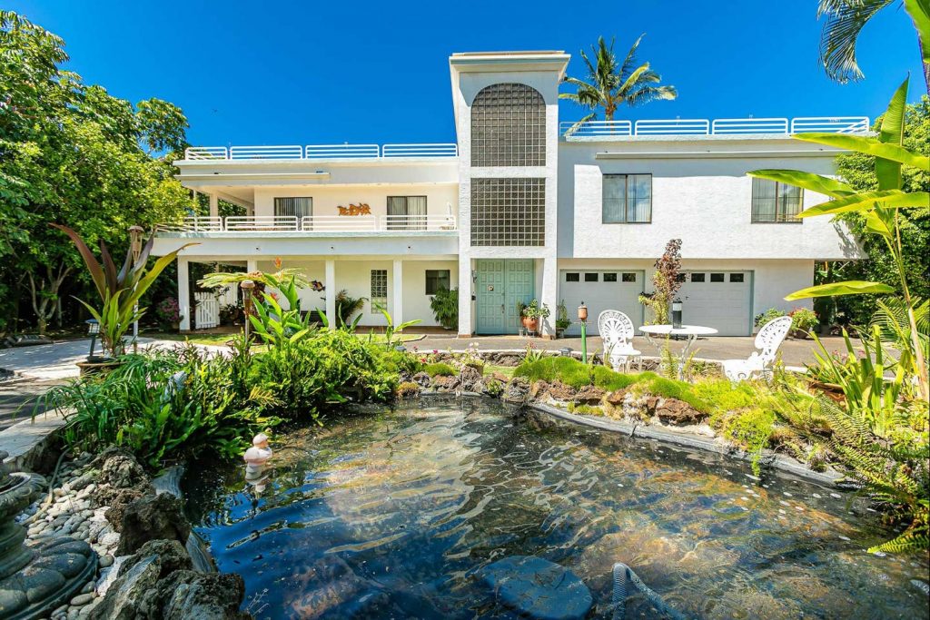A spacious two-story building features large windows and a prominent arch, surrounded by lush tropical greenery. A serene pond, adorned with plants and stones, is situated in the foreground, complemented by outdoor seating on the patio, inviting relaxation amidst nature.