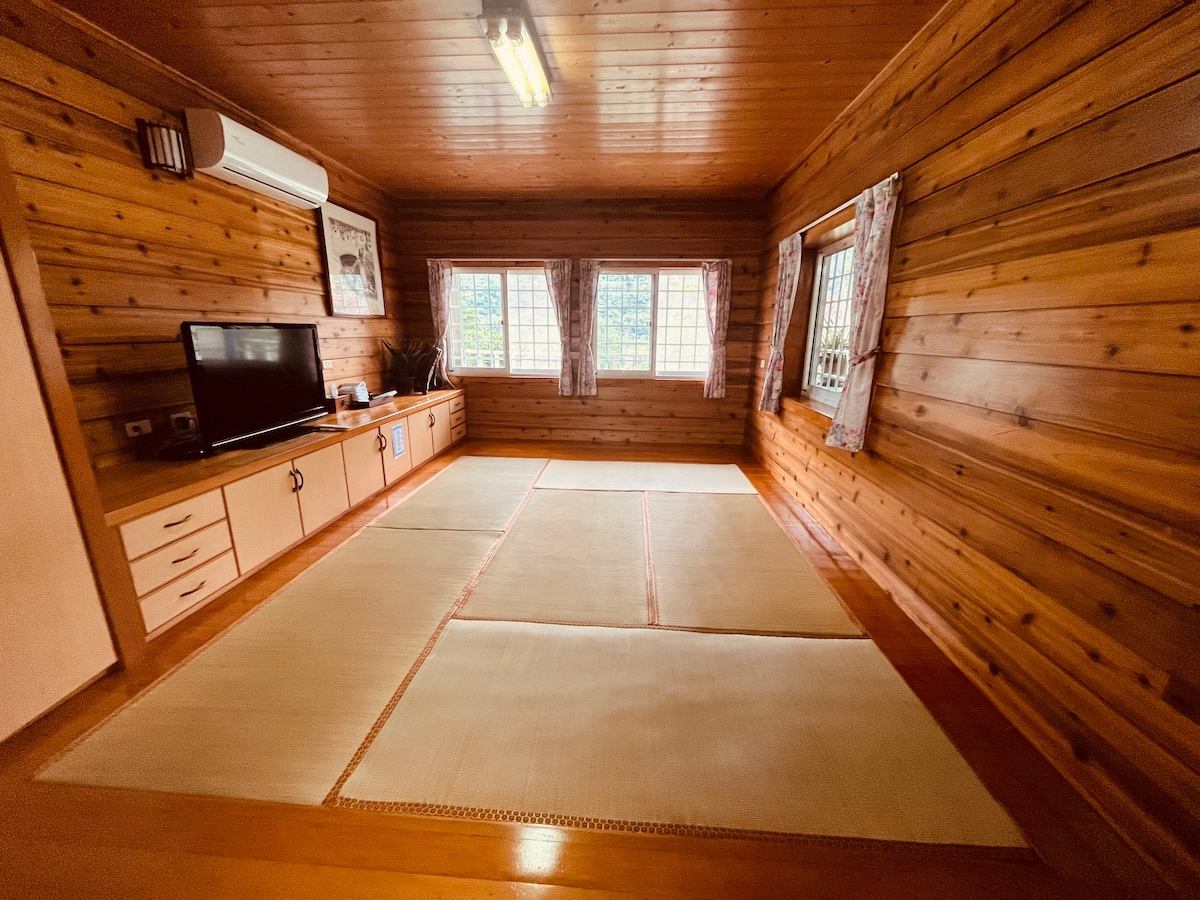 A spacious room features warm wooden walls and flooring, creating a cozy ambiance. Soft, light-colored tatami mats are arranged on the floor. Large windows with decorative curtains allow natural light to fill the area, offering a bright and inviting space.