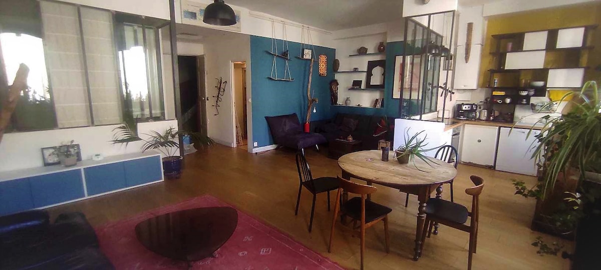 The spacious living area features wooden flooring and a large circular table surrounded by sleek chairs. A dark coffee table complements a plush seating arrangement, including a cozy couch and armchair. The kitchen area is visible, showcasing modern appliances and decorative elements, with plants adding vibrancy.
