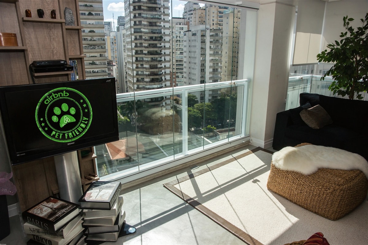 A sunlit living area features a cozy seating arrangement with a large, dark sofa and a woven pouf. A plush throw lies on a light rug, while a TV sits against the wall. Floor-to-ceiling windows offer a view of the surrounding cityscape.