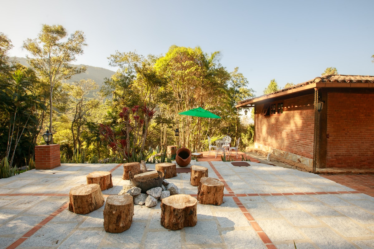 A serene outdoor gathering space features natural wooden logs arranged around a stone fire pit, set on a tiled patio. Lush trees and greenery create a tranquil backdrop, while subtle outdoor furniture, including a shaded area with an umbrella, invites relaxation and connection with nature.