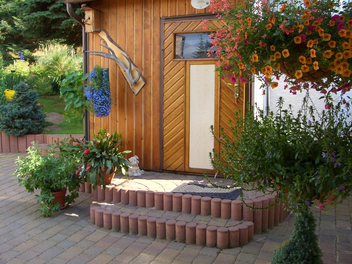 The entrance of a wooden house is framed by vibrant flower pots and lush greenery. A wooden door features a decorative panel, while clay bricks create a tiered step leading up to the entrance. Hanging plants add color and charm to the welcoming space.