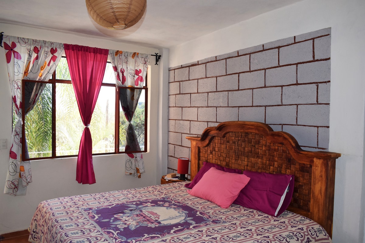 A bright bedroom features a king-size bed adorned with a patterned blanket and colorful throw pillows. Large windows allow natural light to fill the space, while floral curtains frame the view of the greenery outside. A decorative woven lampshade hangs from the ceiling.